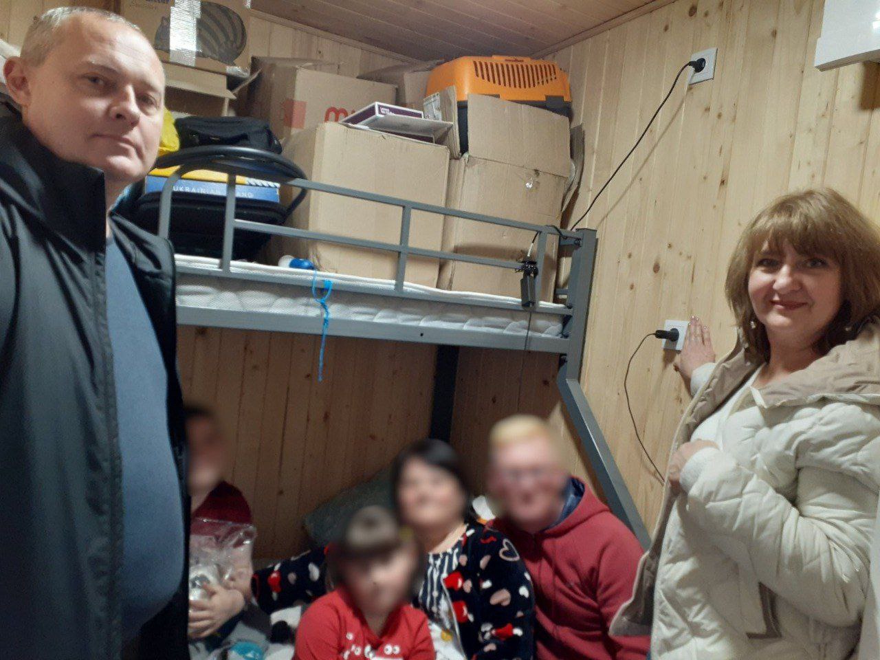 Чоловік і жінка стоять у приміщенні з дерев'яними стінами поруч із двоярусним ліжком, на якому сидять розмиті фігури. На верхньому ярусі ліжку та навколо нього багато картонних коробок.