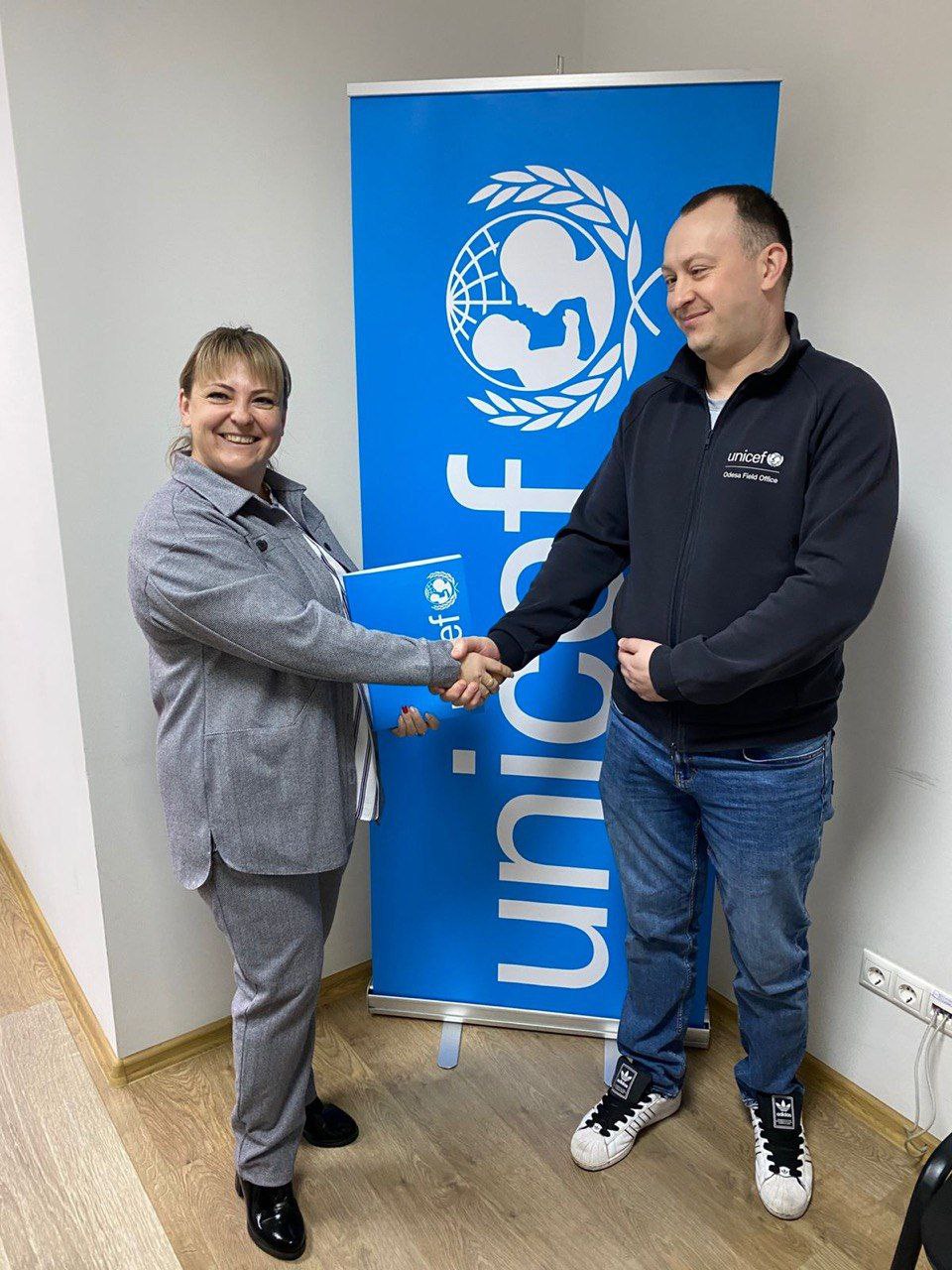 Двоє людей — чоловік і жінка — потискають один одному руки на тлі великого синього банера ЮНІСЕФ (UNICEF). Жінка, одягнена у сірий брючний костюм, посміхається і тримає синю теку. Чоловік, одягнений у чорну толстовку з логотипом ЮНІСЕФ, також усміхається.