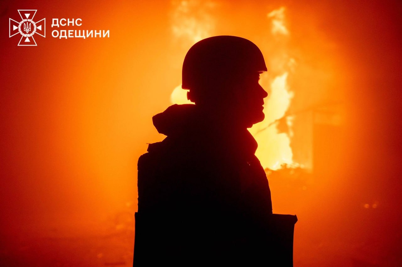 Силует рятувальника в касці, знятий крупним планом на тлі яскраво-оранжевого полум'я, яке охопило будівлю. Язики вогню створюють інтенсивне світіння, що підкреслює силует. У лівому верхньому куті розташований логотип ДСНС Одещини.