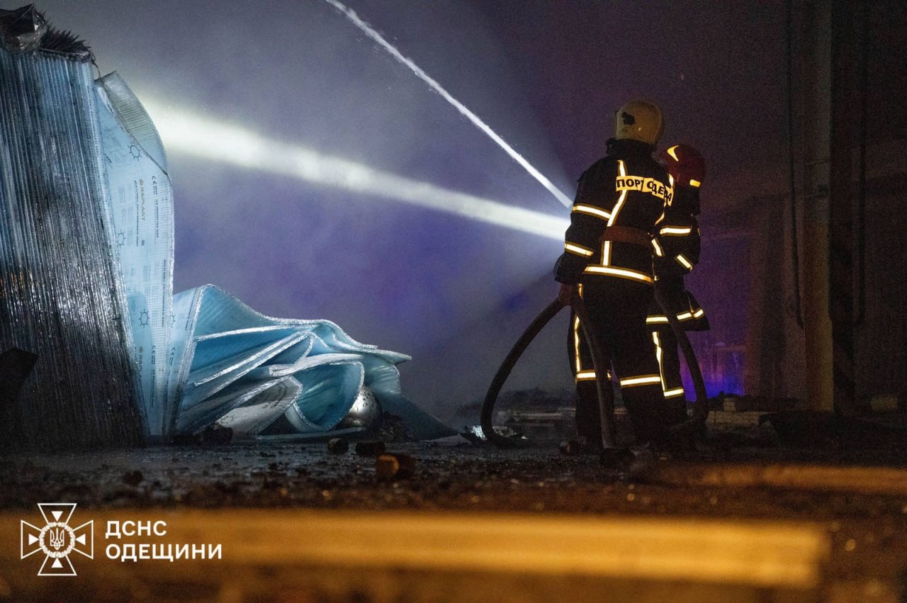 Двоє пожежників у повній формі з написом: ПОРЯТУЄМО на спині стоять у темряві та поливають водою вогнище. Вони стоять біля великої купи синьо-білого матеріалу (можливо, утеплювача або зруйнованих конструкцій). Світло від ліхтаря яскраво освітлює стовп води та частину матеріалу. У лівому нижньому куті розташований логотип ДСНС Одещини.