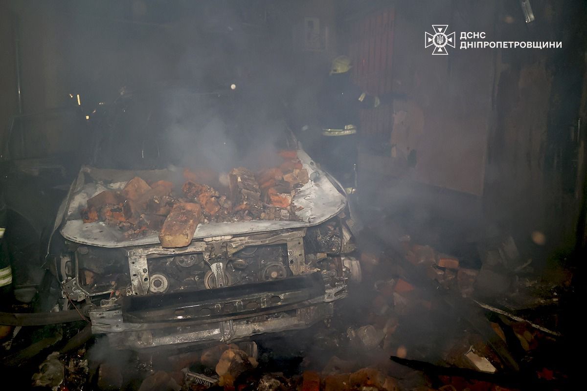 Згорілий, сильно пошкоджений автомобіль, накритий купою уламків цегли, стоїть у темному гаражі або приміщенні. Автомобіль, ймовірно, був знищений під час пожежі, від нього йде густий дим. Праворуч, на задньому плані, стоїть рятувальник у захисному шоломі. У правому верхньому куті розташований логотип ДСНС Дніпропетровщини.