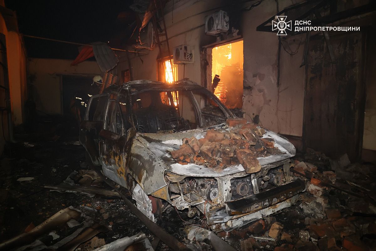 Згорілий, сильно пошкоджений автомобіль, накритий купою уламків цегли, стоїть у темному приміщенні. Праворуч у дверному отворі видно яскраве полум'я. У кадрі присутній рятувальник, а вся сцена оповита димом. У правому верхньому куті розташований логотип ДСНС Дніпропетровщини.