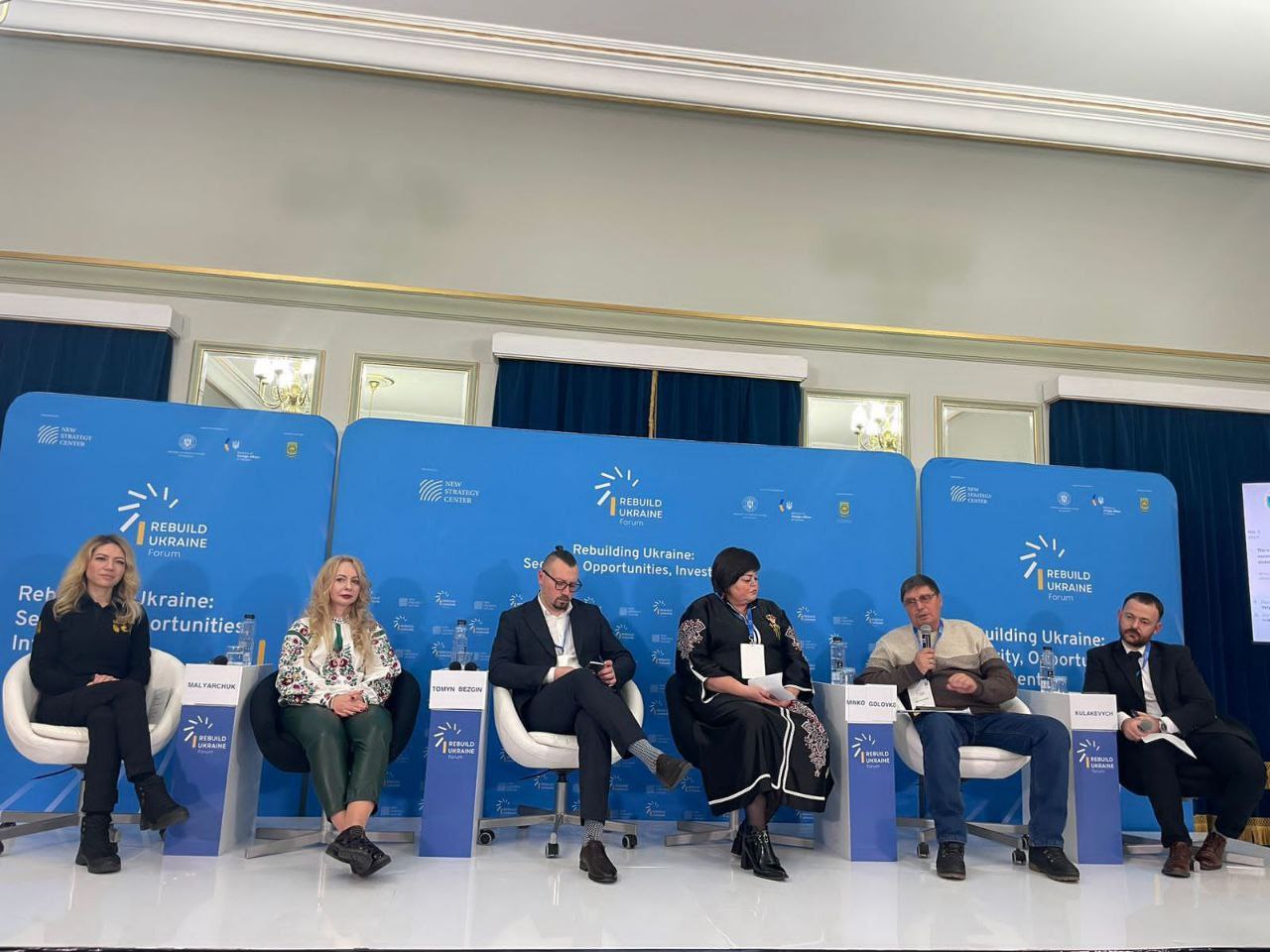 Панельна дискусія на форумі REBUILD UKRAINE з шістьма учасниками, які сидять на стільцях перед синіми банерами.