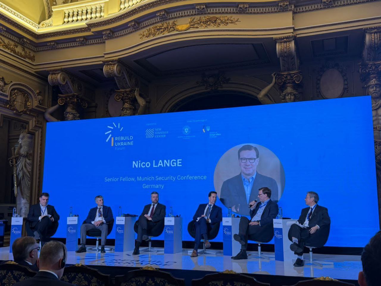 Панельна дискусія в золотистому бароковому залі, де шестеро чоловіків сидять на сцені перед великим синім екраном з логотипом REBUILD UKRAINE FORUM та інформацією про учасника на онлайн-зв'язку.