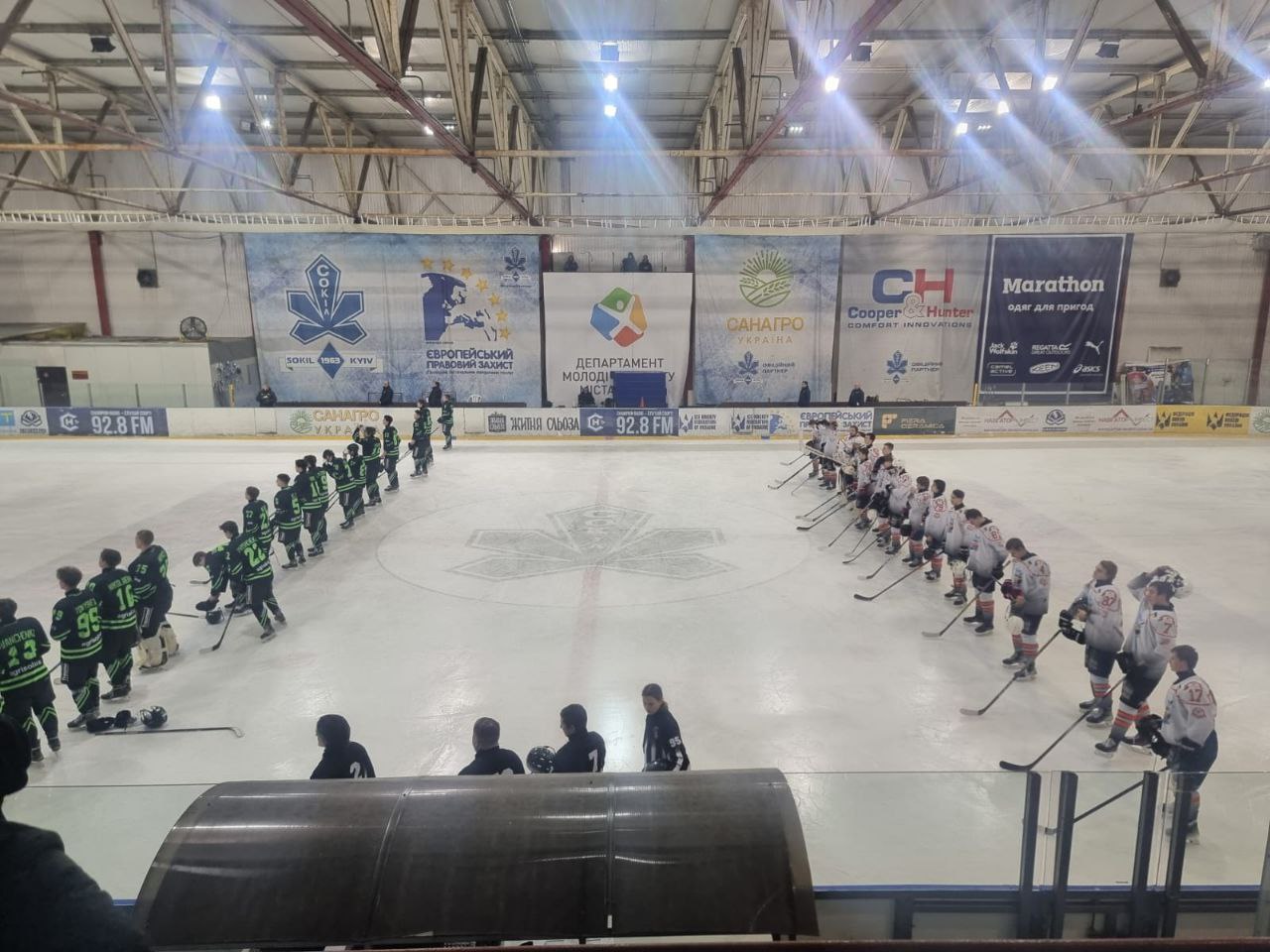 Дві команди хокеїстів (у чорно-зеленій та помаранчево-білій формі) шикуються на льоду обличчям одна до одної перед початком або після закінчення матчу. На задньому плані видно рекламні банери під дахом арени.