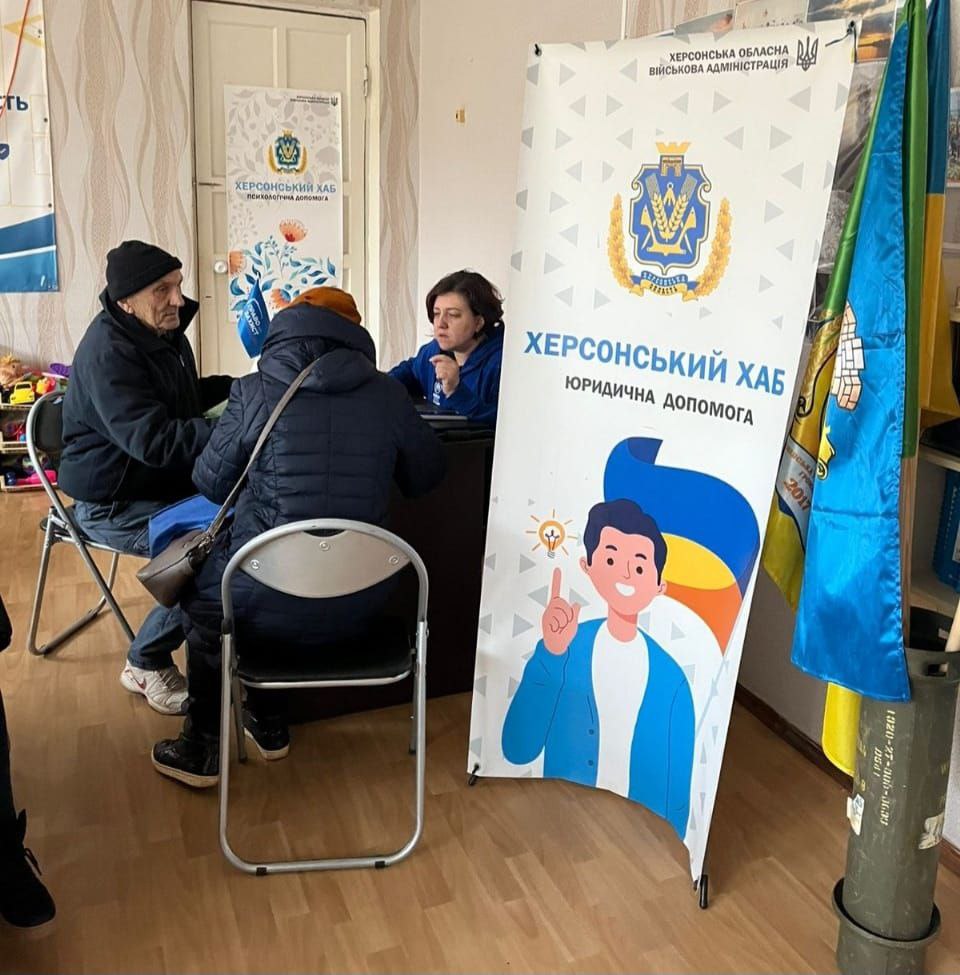 Два літні відвідувачі отримують консультацію від жінки-представниці за столом. Поруч стоїть великий банер ХЕРСОНСЬКИЙ ХАБ ЮРИДИЧНА ДОПОМОГА від Херсонської обласної військової адміністрації, а також видно прапори України.