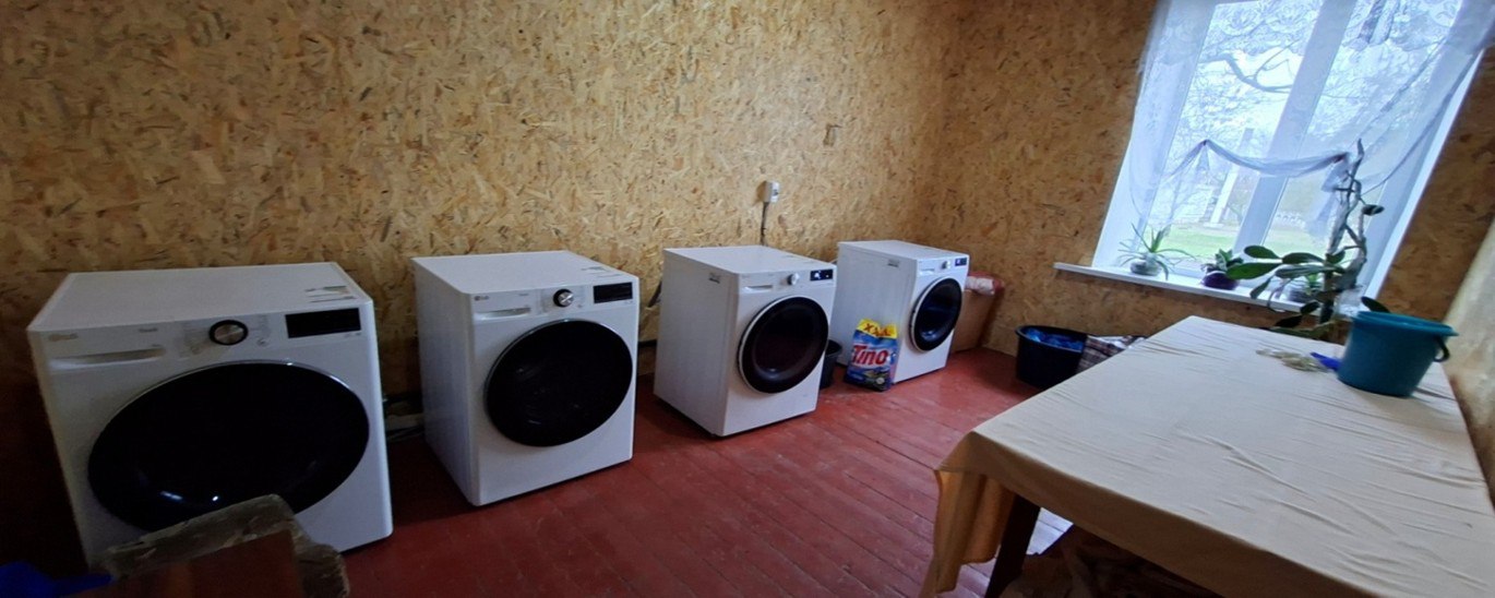 Чотири білі пральні машини стоять уздовж стіни у кімнаті з дерев'яними панелями та червоною підлогою; праворуч видно стіл і вікно.