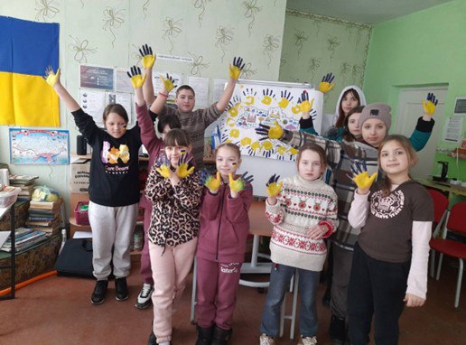 Група дітей стоїть у класі, піднявши руки, пофарбовані у синьо-жовті кольори; на стіні ліворуч висить український прапор.