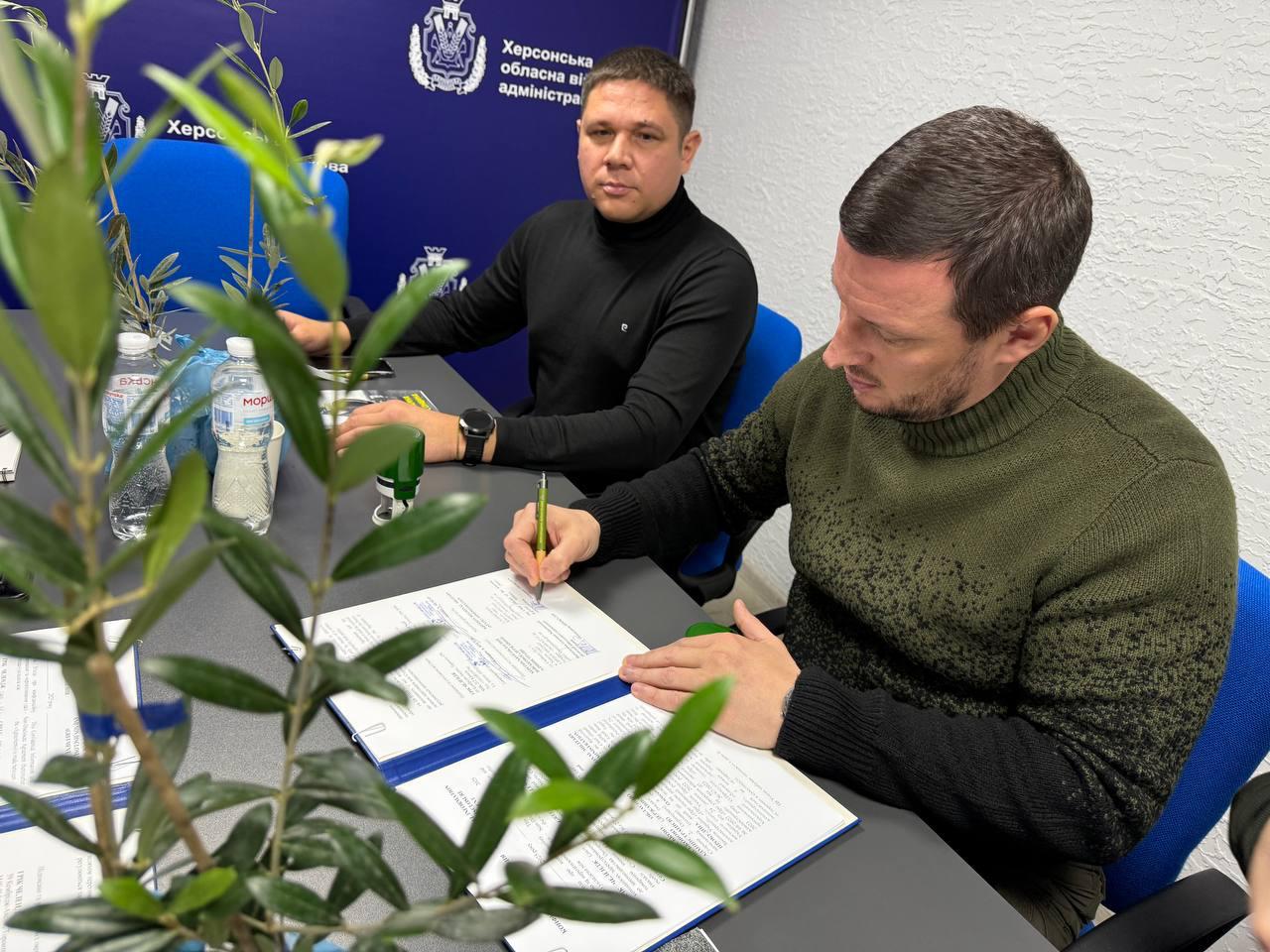 Двоє чоловіків сидять за столом, один із них (у світшоті кольору хакі) підписує документи, а інший (у чорній водолазці) дивиться на нього; на столі видно рослину та пляшки з водою, а на задньому плані — герб Херсонської обласної військової адміністрації.