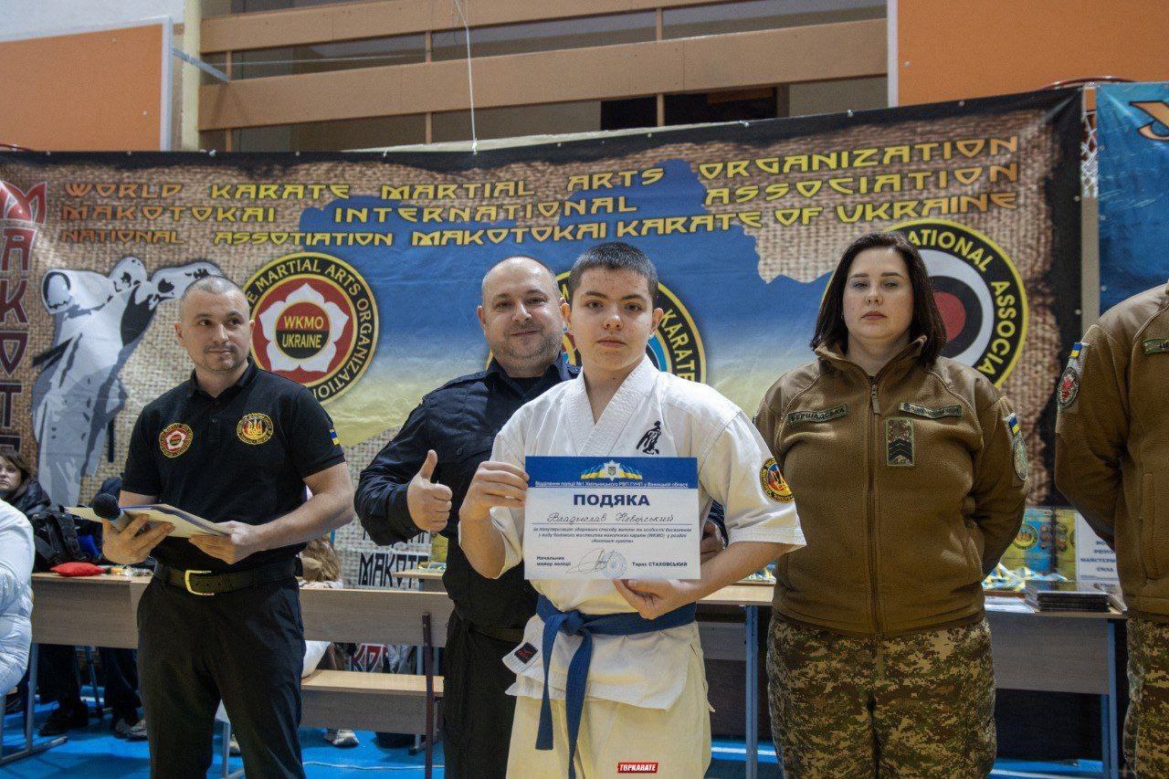 Хлопець у білому кімоно з синім поясом стоїть по центру і тримає сертифікат, показуючи великий палець вгору. Його обіймає чоловік зліва. Праворуч від нього стоїть жінка у військовій формі. На задньому плані банер з написами: WORLD KARATE MARTIAL ARTS ORGANIZATION ASSOCIATION INTERNATIONAL MAKOTOKAI KARATE OF UKRAINE.