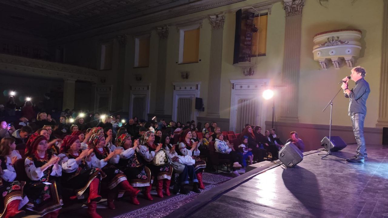 Виконавець стоїть на сцені в залі, тримаючи мікрофон і співаючи. На сцену спрямовано яскраве світло. Глядачі, включно з групою жінок у традиційних українських вишитих костюмах (з червоними чобітьми та хустками), сидять у залі, багато хто з них аплодує або знімає подію на телефони.