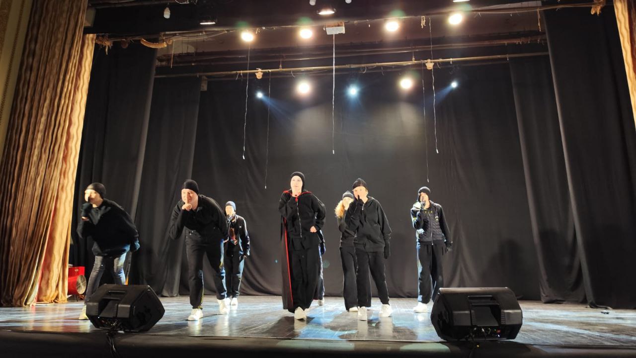 Група з семи підлітків у чорному танцювальному одязі та шапках виступає на сцені, виконуючи динамічний рух. Одна людина по центру одягнена в чорний плащ із червоними елементами.