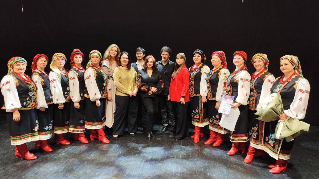 Група з приблизно 16 людей, більшість з яких жінки, стоять на сцені на тлі чорної завіси. Жінки переважно одягнені у традиційні українські вишивані костюми з червоними чобітьми та хустками. У центрі стоять декілька людей у сучасному одязі, включно з двома чоловіками. Деякі жінки тримають нагороду та букет квітів.