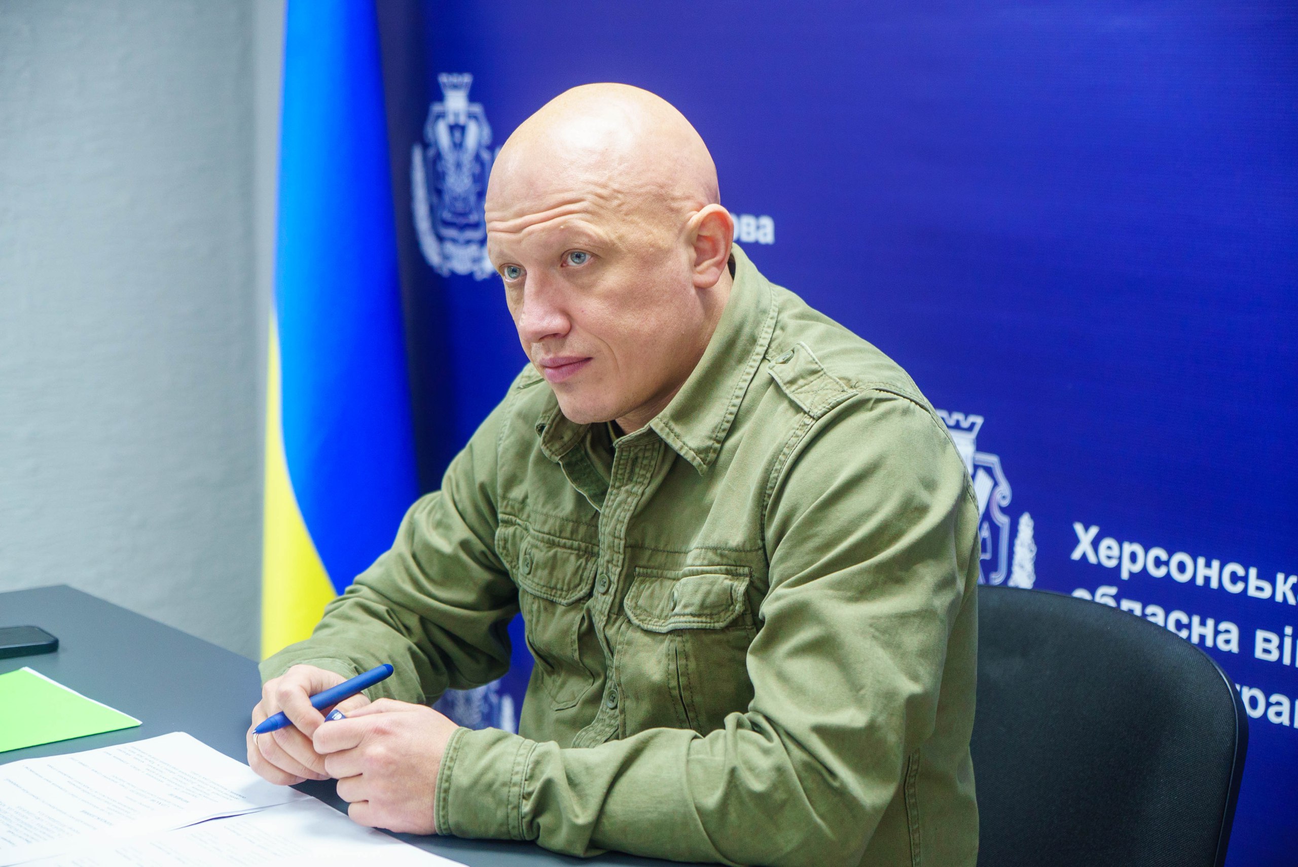 Портретний знімок: чоловік із поголеною головою у військовій сорочці кольору хакі сидить за столом, тримаючи ручку, на фоні синього банера Херсонська обласна військова адміністрація та прапорів.