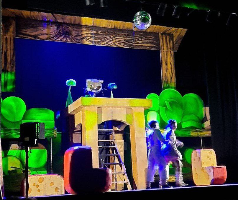 Сцена з театральної вистави з яскравими декораціями. У центрі – жовта конструкція-будинок зі сходами, на якій стоїть ляльковий персонаж-тварина. Сцена освітлена синіми та зеленими кольорами, а по боках – великі декорації у вигляді шматків сиру.