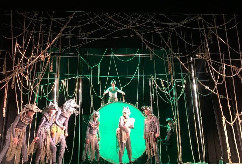 Сцена з театральної вистави, освітлена зеленим світлом. На сцені – актори в сірих костюмах із масками тварин (вовків). Декорації складаються з великої кількості мотузок, що звисають, і великого круглого зеленого екрана по центру. Один актор стоїть на підвищенні позаду кола.