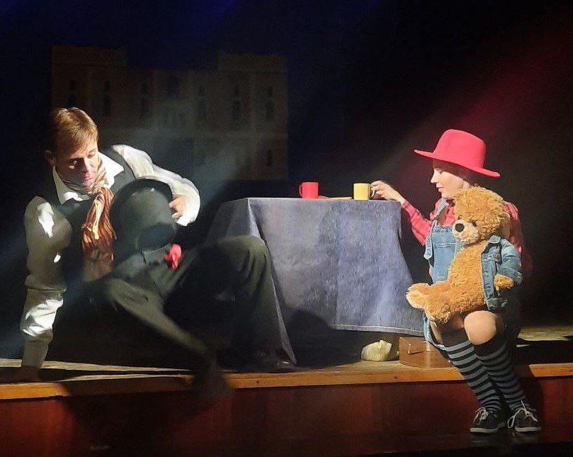 Два актори на сцені під час вистави. Чоловік у світлій сорочці та темних штанах сидить біля столу, тримаючи капелюх. Жінка в рожевому капелюсі, джинсовому комбінезоні та смугастих гольфах сидить на краю сцени, обіймаючи великого плюшевого ведмедика.