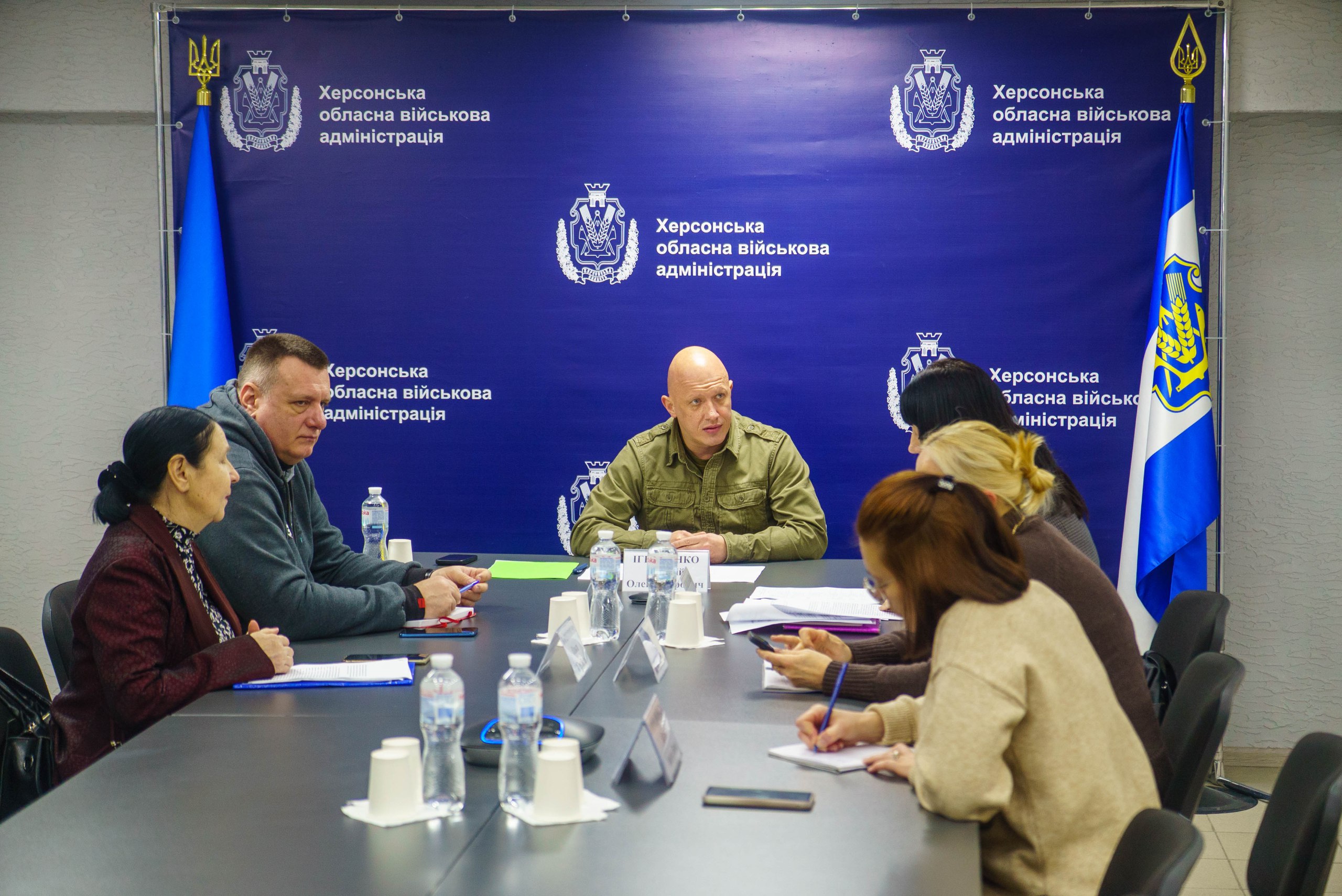 Група людей сидить за великим столом під час наради, очолюваної чоловіком у військовій формі на фоні банера Херсонська обласна військова адміністрація з прапорами.