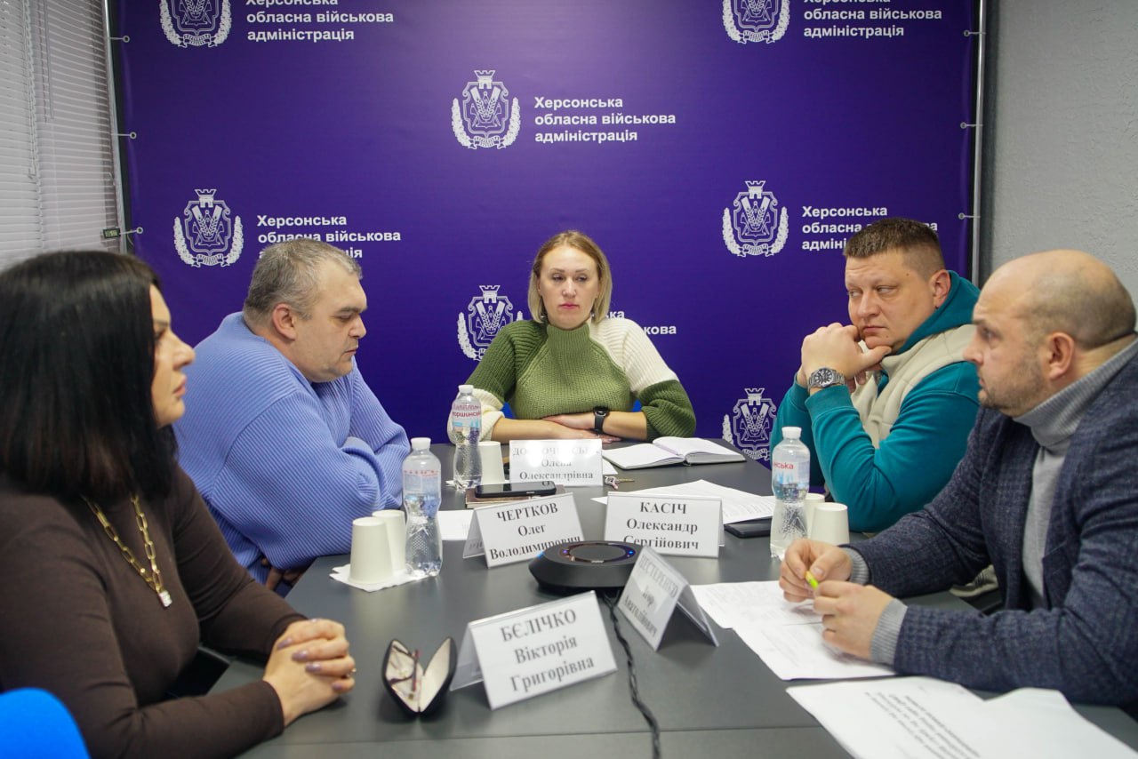 Група з п'яти осіб, четверо чоловіків та одна жінка, сидить за столом під час зустрічі або наради. На задньому плані — синій фон із гербом і написом: Херсонська обласна військова адміністрація. На столі перед учасниками розташовані таблички з прізвищами.