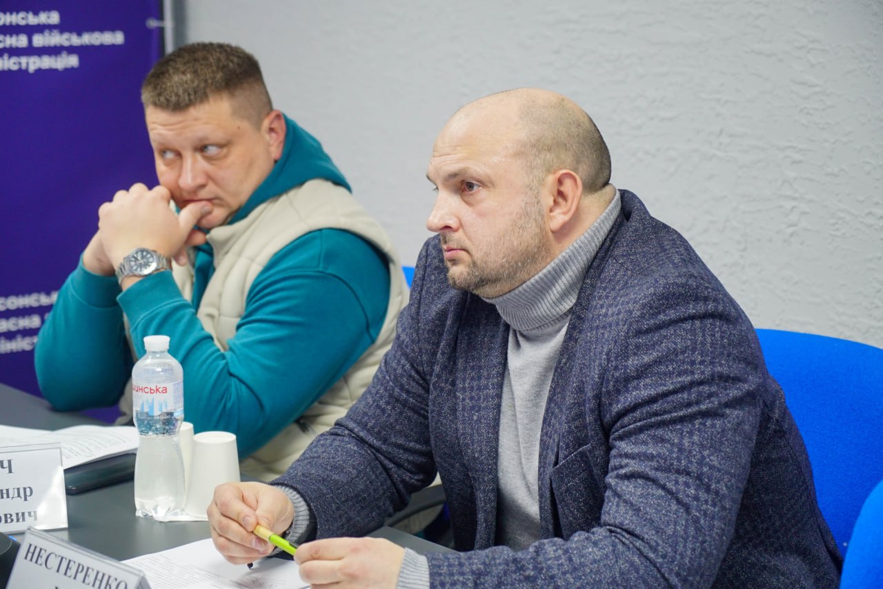 Двоє чоловіків сидять за столом під час зустрічі. На передньому плані — лисий чоловік у темно-синьому піджаку та сірому гольфі, який дивиться вбік. Позаду нього сидить інший чоловік у білій жилетці та бірюзовій толстовці, який підпер рукою підборіддя, виглядаючи зосередженим.