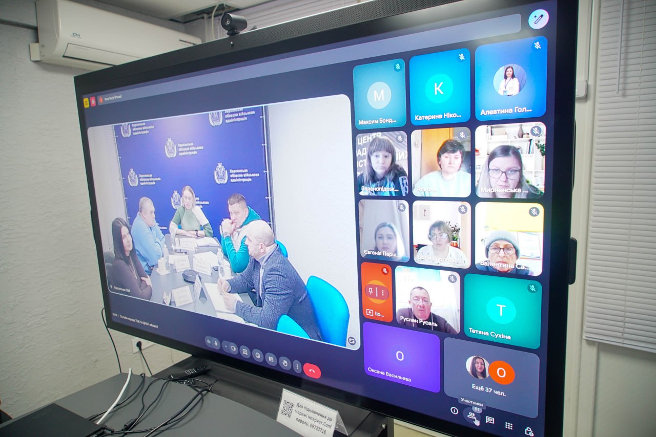 Великий екран, що демонструє відеоконференцію в додатку для онлайн-зустрічей. На головному екрані зліва показано п’ятьох учасників, які сидять за столом, а праворуч — галерея з вікнами інших учасників, деякі з яких мають лише ініціали. На знімку екрана конференції видно фрагмент зустрічі в Херсонській обласній військовій адміністрації.