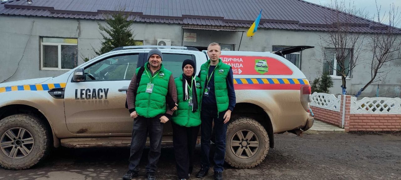 Троє медиків-волонтерів у зелених жилетах стоять біля брудної вантажівки з написом: LEGACY та МЕДИЧНА КОМАНДА на фоні будівлі з прапором України.