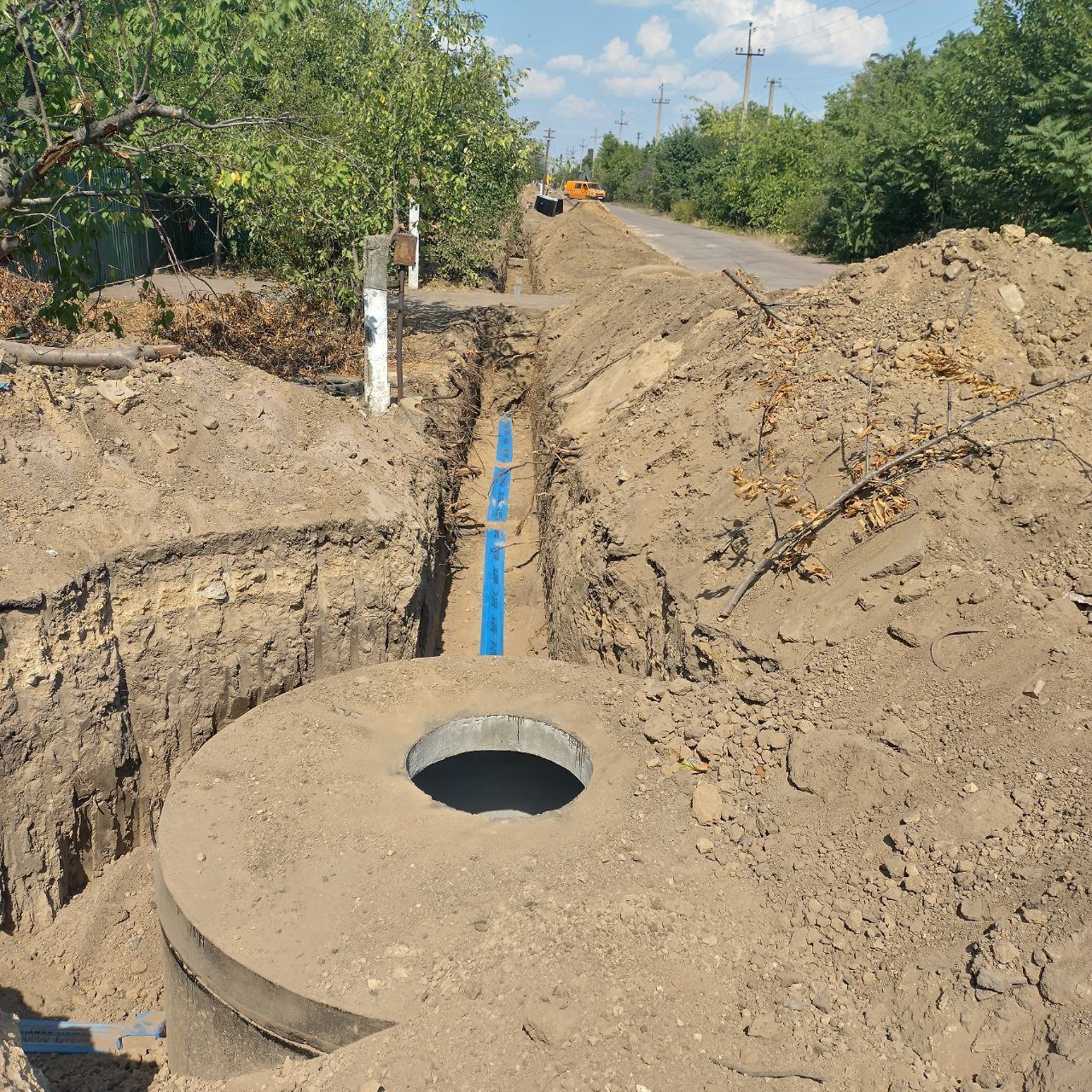 Процес прокладання підземних комунікацій уздовж сільської дороги. На передньому плані видно відкритий бетонний колодязь у глибокій траншеї, в якій прокладена синя труба. Обабіч траншеї насипані купи землі, а вдалині видніється спецтехніка та зелені дерева.