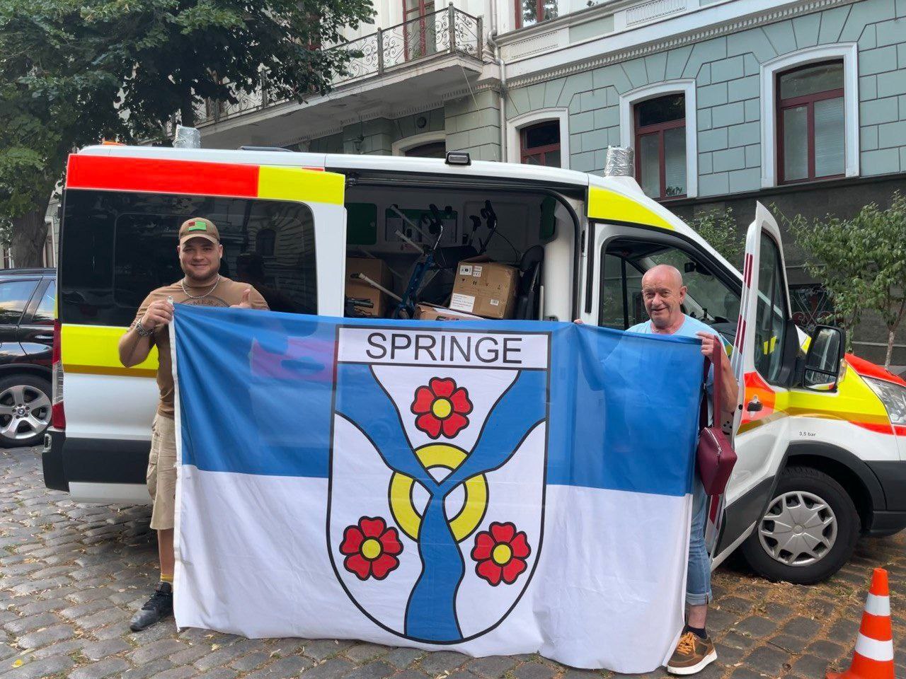 Двоє чоловіків стоять поруч із білим мікроавтобусом і тримають великий біло-синій прапор міста Шпрінге (Springe) з гербом. Автомобіль завантажений коробками та медичним обладнанням, що вказує на доставку гуманітарної допомоги.