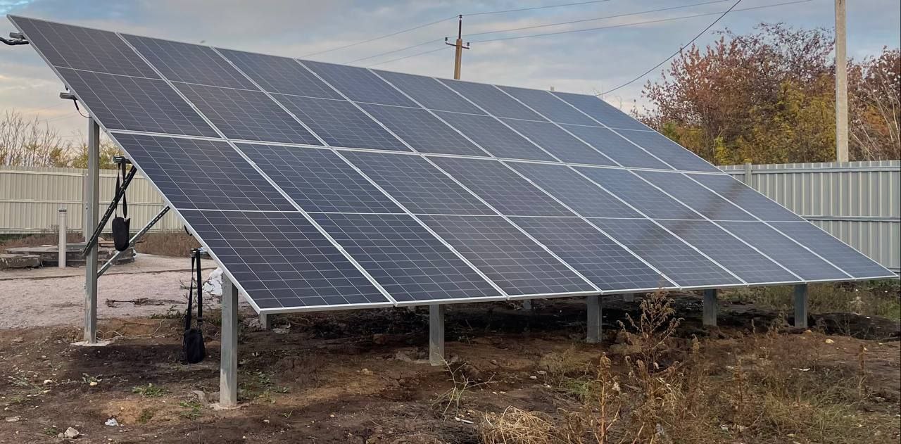 Наземна сонячна електростанція, що складається з кількох рядів фотоелектричних панелей, встановлених на металевих опорах. Установка розміщена на відкритій ділянці поруч із парканом, на фоні осінніх дерев та хмарного неба.