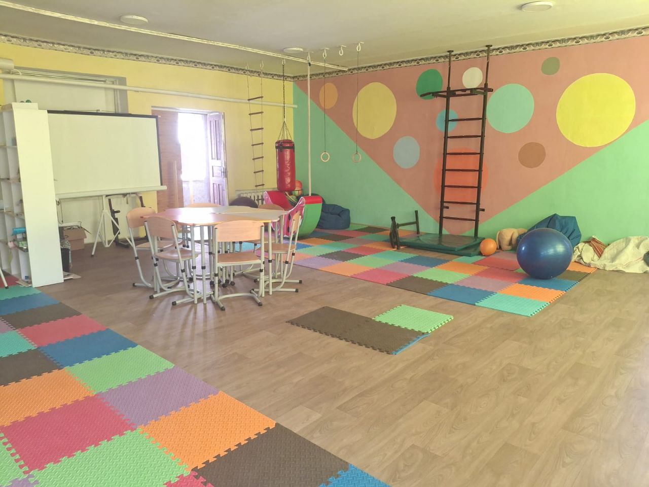 Дитяча ігрова та навчальна кімната з яскравим дизайном. У приміщенні розташована зона для занять зі столами та стільцями, а також спортивний куточок із драбиною, боксерською грушею та фітболом. Підлога застелена різнокольоровим м'яким покриттям-пазлом, а стіни прикрашені великими кольоровими колами.