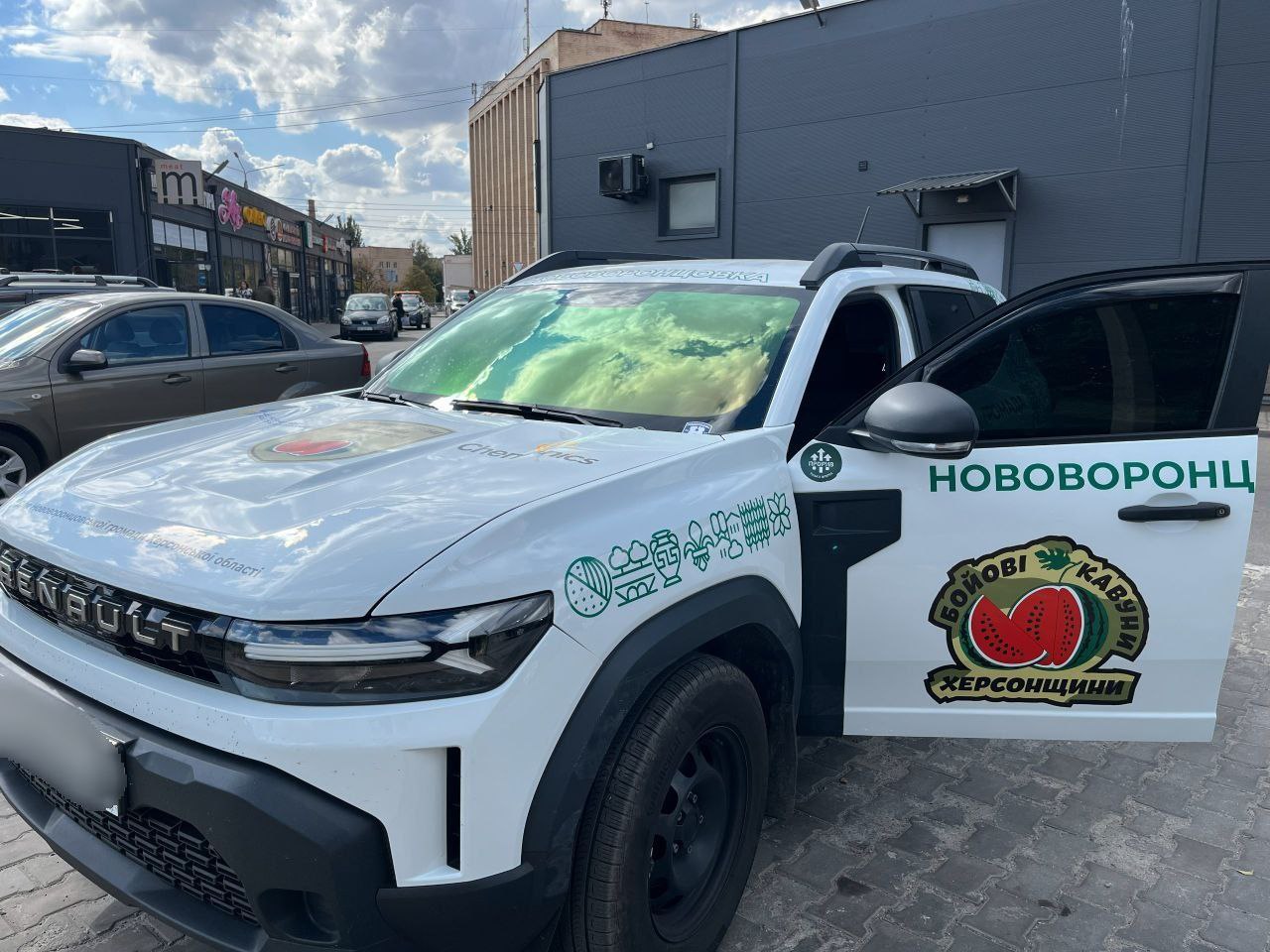 Білий позашляховик Renault з патріотичним брендуванням, припаркований на вулиці. На дверцятах автомобіля зображено логотип із кавуном та написом: Бойові кавуни Херсонщини, а також назву селища Нововоронцовка. На капоті та боках автівки розміщені традиційні українські орнаменти та написи.