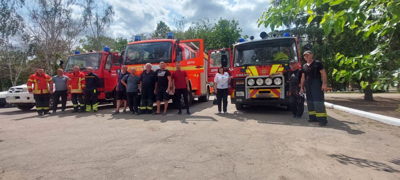 Група рятувальників та цивільних стоїть на фоні трьох червоних пожежних автомобілів під відкритим небом. На бортах машин видно написи ДПО (Добровільна пожежна охорона), а на одній з них — логотип Volvo.