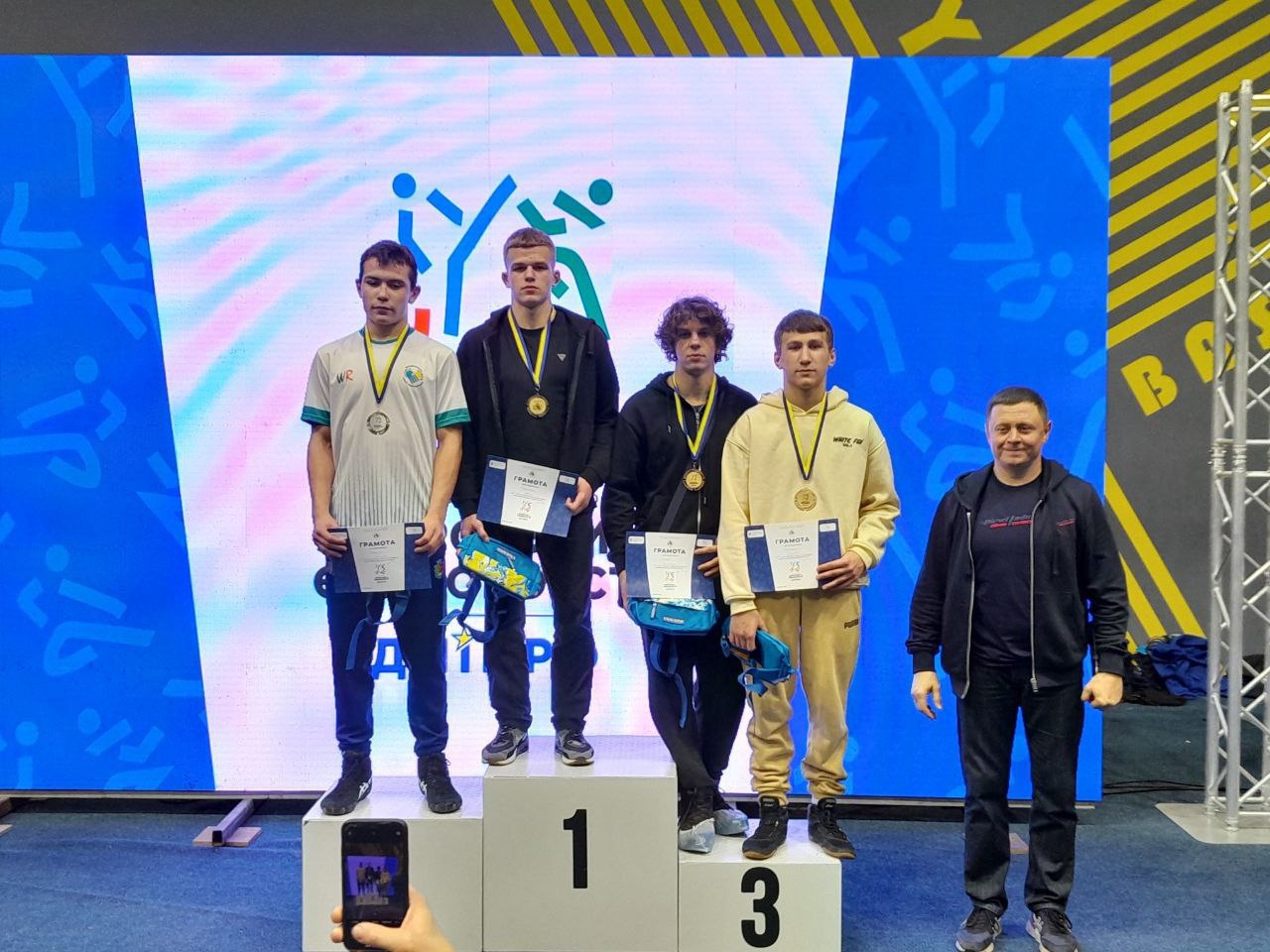 Четверо молодих спортсменів стоять на подіумі для нагородження (1, 3, 2 місця) з медалями та грамотами поруч із чоловіком у чорній куртці, на фоні екрана з логотипом спортивного турніру.