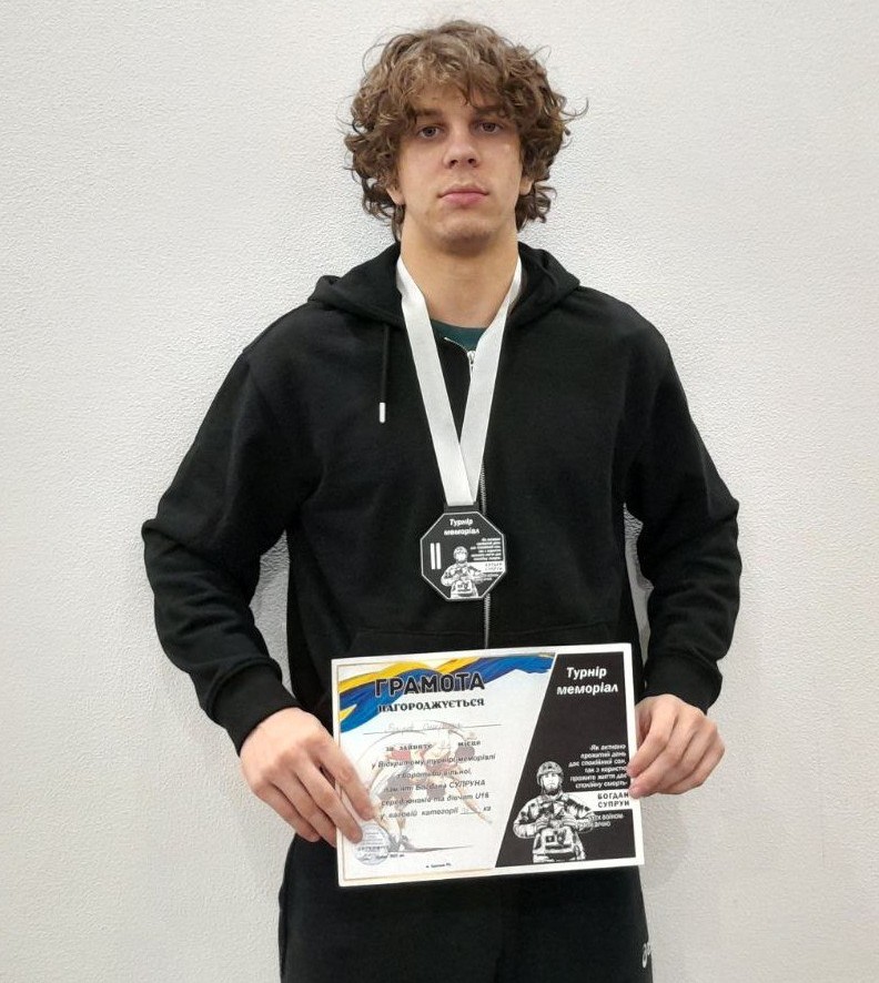 Молодий чоловік з кучерявим волоссям у чорному худі стоїть, тримаючи Грамоту та медаль, які він отримав на турнірах вільної боротьби.