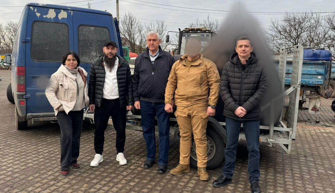 П'ять людей – жінка та четверо чоловіків (один у військовій формі) – стоять перед синім мікроавтобусом та причепом у дворі.