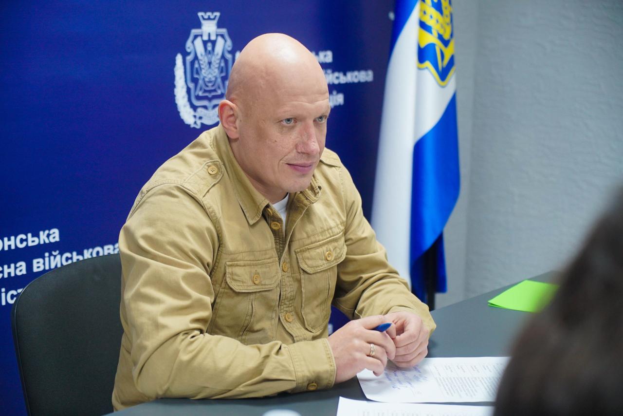 Крупний план лисого чоловіка в пісочній сорочці військового стилю, який сидить за столом і посміхається. За ним видно темно-синій фон з текстом: Херсонська обласна військова адміністрація та жовто-блакитний прапор.