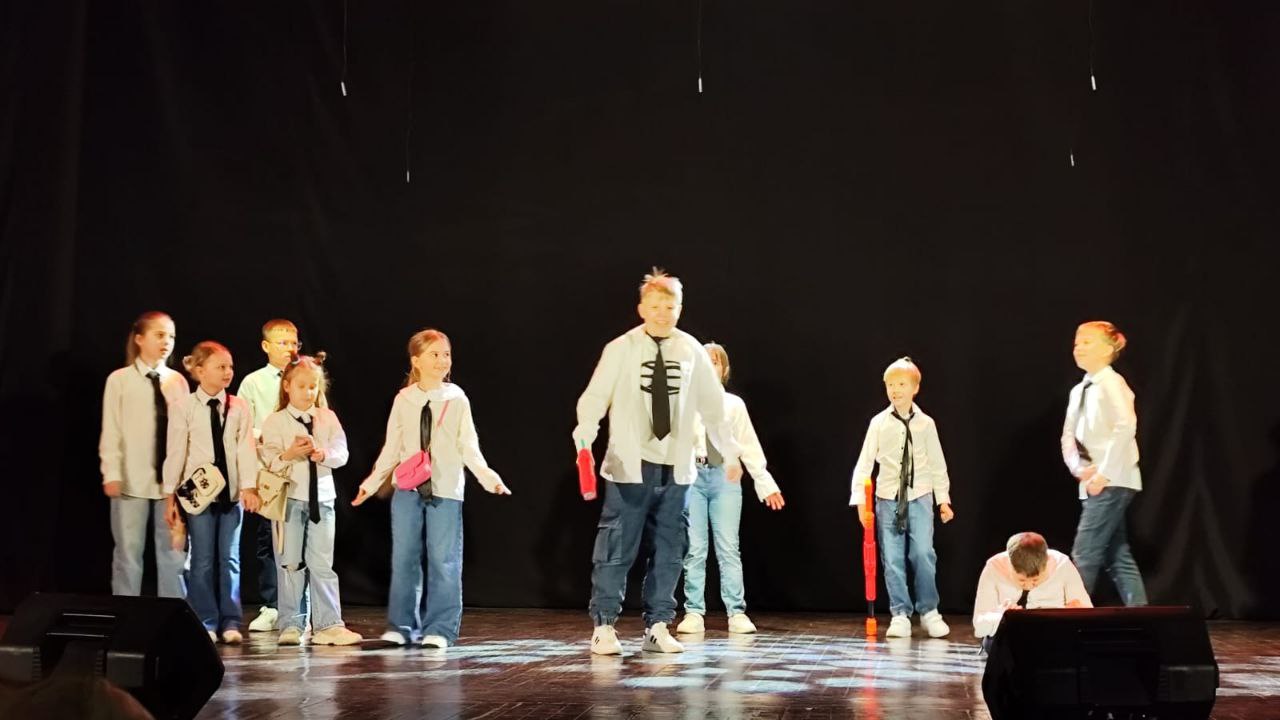 Деcять дітей стоять на сцені перед чорним тлом, освітлені прожекторами. Вони одягнені в білі сорочки, чорні краватки та сині джинси. Хлопчик у центрі стоїть спереду, а один хлопчик нахилився до сцени на передньому плані. Це схоже на танцювальний чи театральний виступ.
