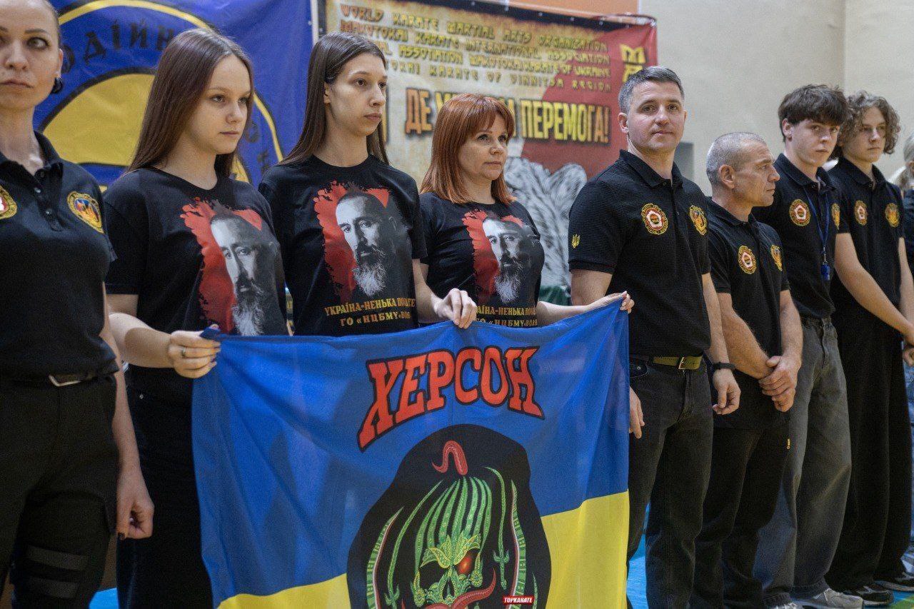 Група з восьми людей, одягнених у чорні футболки (три з яких мають на принті портрети двох чоловіків і написи), стоїть у ряд. Троє жінок попереду тримають великий синьо-жовтий прапор з написом: ХЕРСОН червоними літерами та зображенням черепа знизу. На задньому плані видно частину банера.