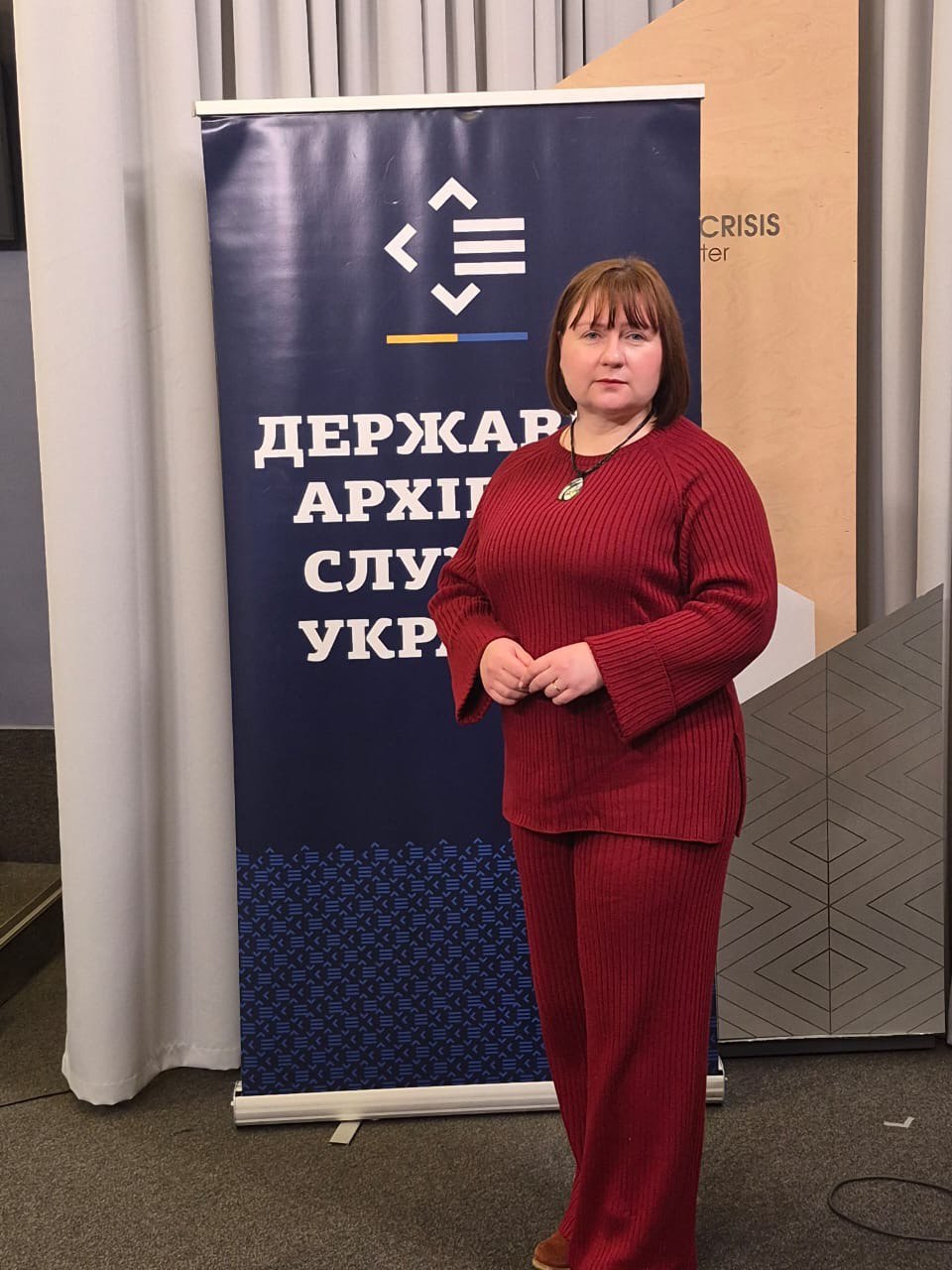 Жінка в червоному костюмі стоїть поруч із банером Державної архівної служби України в приміщенні Ukraine Crisis Media Center.