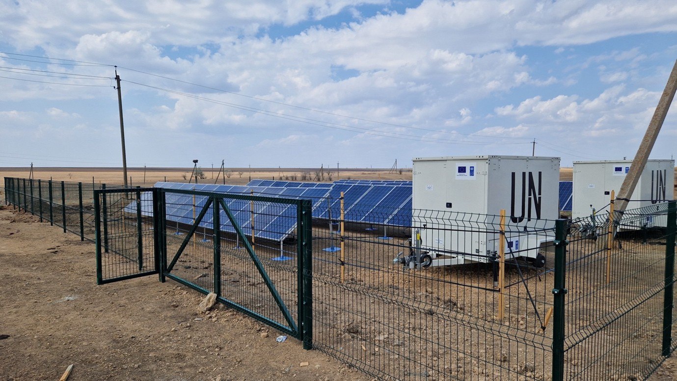 Сонячна електростанція в польових умовах, огороджена парканом. На території встановлено ряди сонячних панелей та два білі мобільні контейнери з маркуванням ООН (UN) та прапором ЄС.