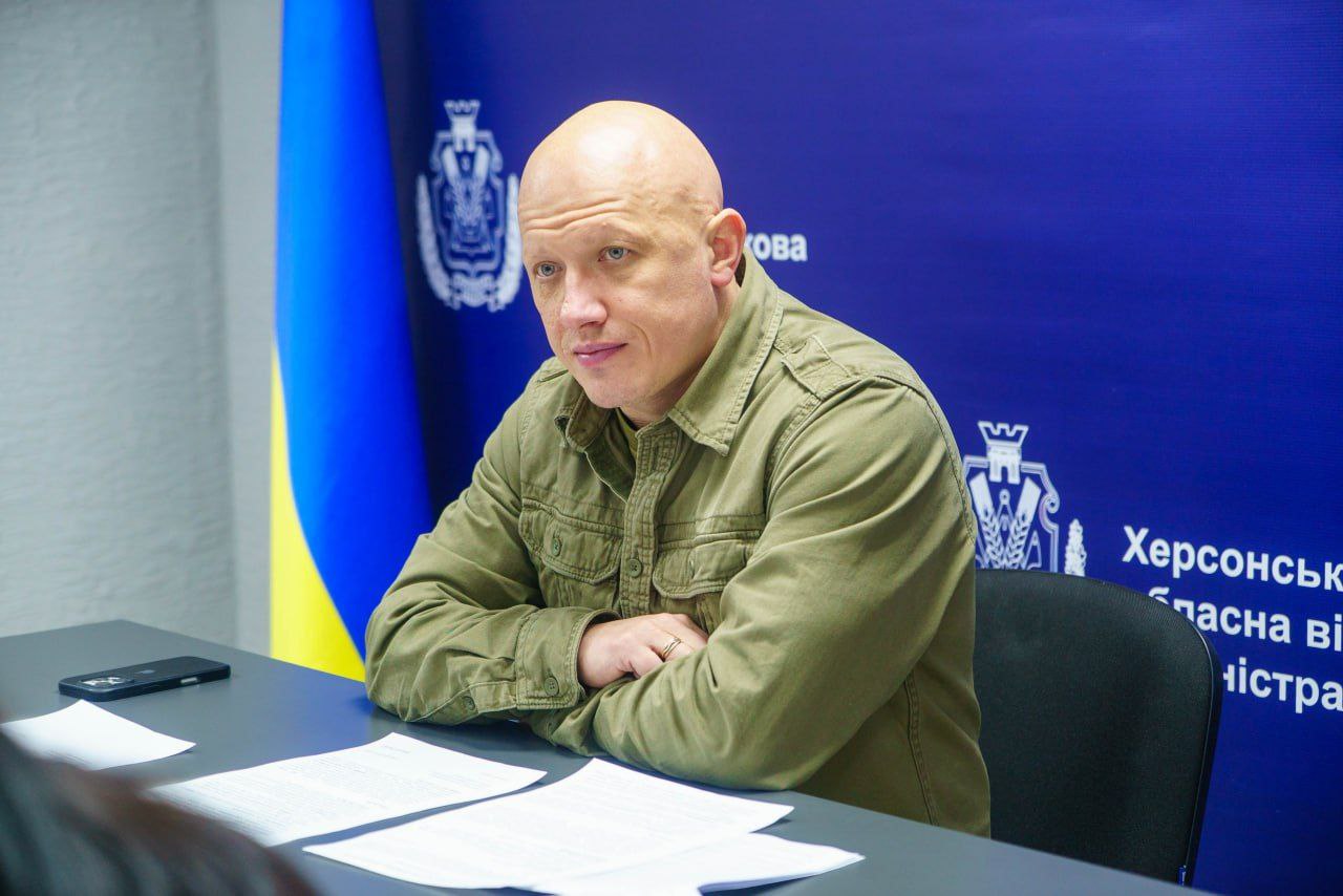 Портрет чоловіка у військовій сорочці кольору хакі, який сидить за столом на фоні синього банера з логотипом та написом: Херсонська обласна військова адміністрація.