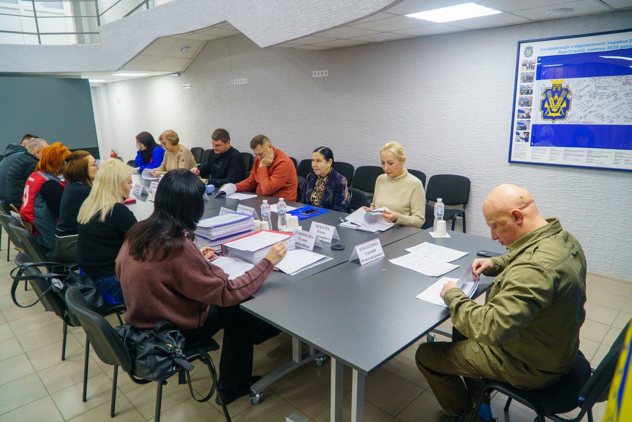 Група людей сидить за довгим робочим столом під час наради, розглядаючи документи. На передньому плані праворуч — чоловік у військовій сорочці кольору хакі, який вивчає папери.