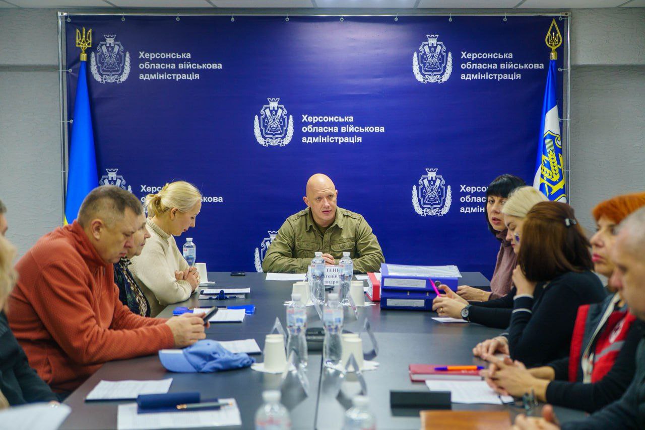 Нарада в Херсонській обласній військовій адміністрації: група посадовців та фахівців за робочим столом під головуванням чоловіка у військовій формі на фоні синього брендованого банера.