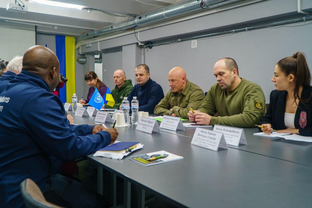 Робоча зустріч представників місцевої влади з представниками міжнародних організацій у сучасному кабінеті. Учасники сидять за довгим сірим столом, на якому розміщені таблички з іменами, пляшки з водою та настільні прапорці України та Організації Об'єднаних Націй.