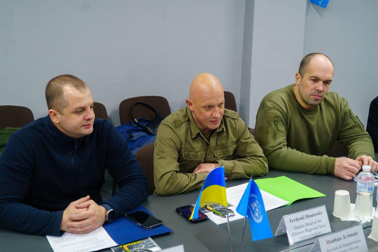 Троє посадовців у цивільному та військовому одязі сидять за столом під час офіційної зустрічі. У центрі — чоловік у військовій сорочці кольору хакі, перед яким встановлено настільні прапорці України та ООН.