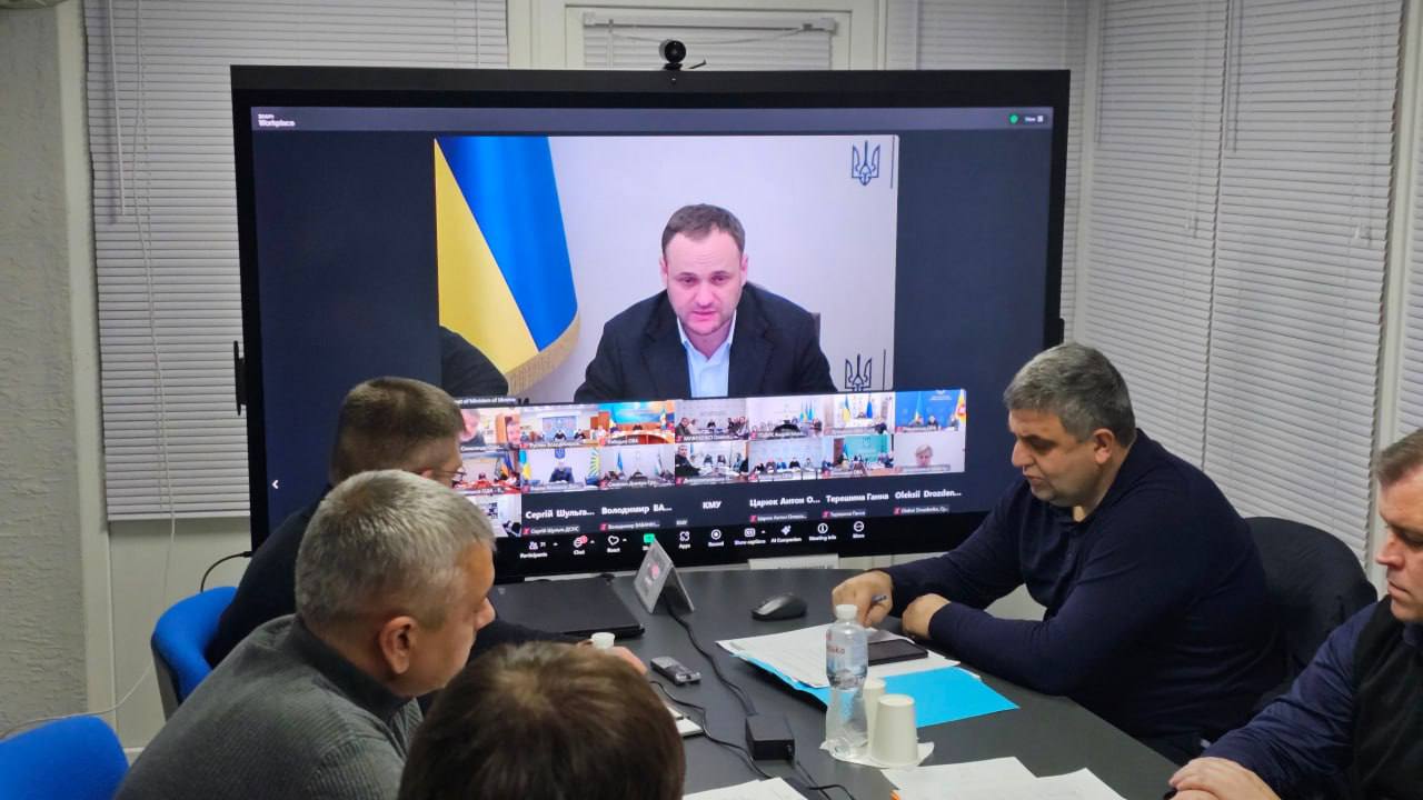 Учасники наради в офісному приміщенні дивляться на великий екран, де триває відеоконференція за участі представника державної влади на фоні прапора України.