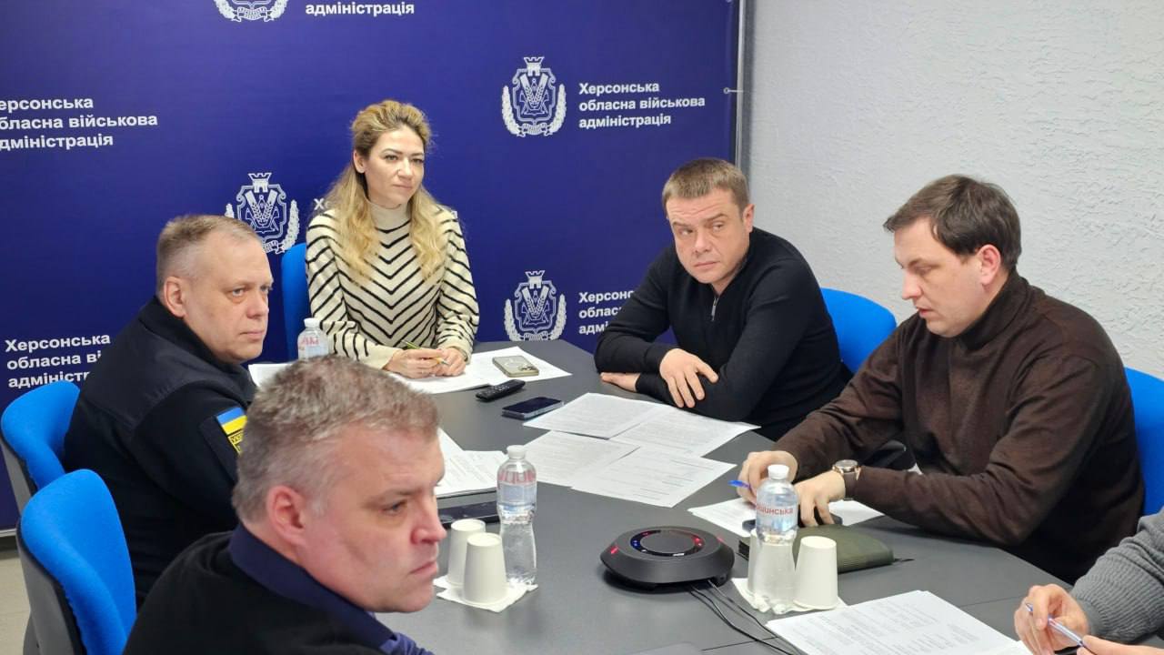Робоча зустріч у Херсонській обласній військовій адміністрації. П'ятеро посадовців сидять за столом під час обговорення на фоні брендованої стіни з гербом області.