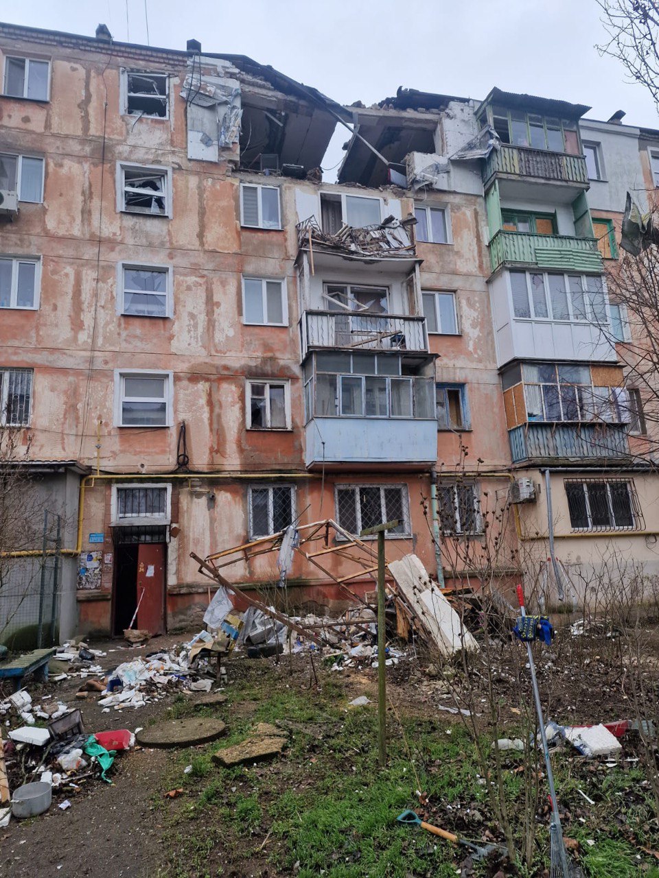 Пошкоджений багатоквартирний будинок з пробитим дахом, зруйнованим балконом і уламками на землі після обстрілу.