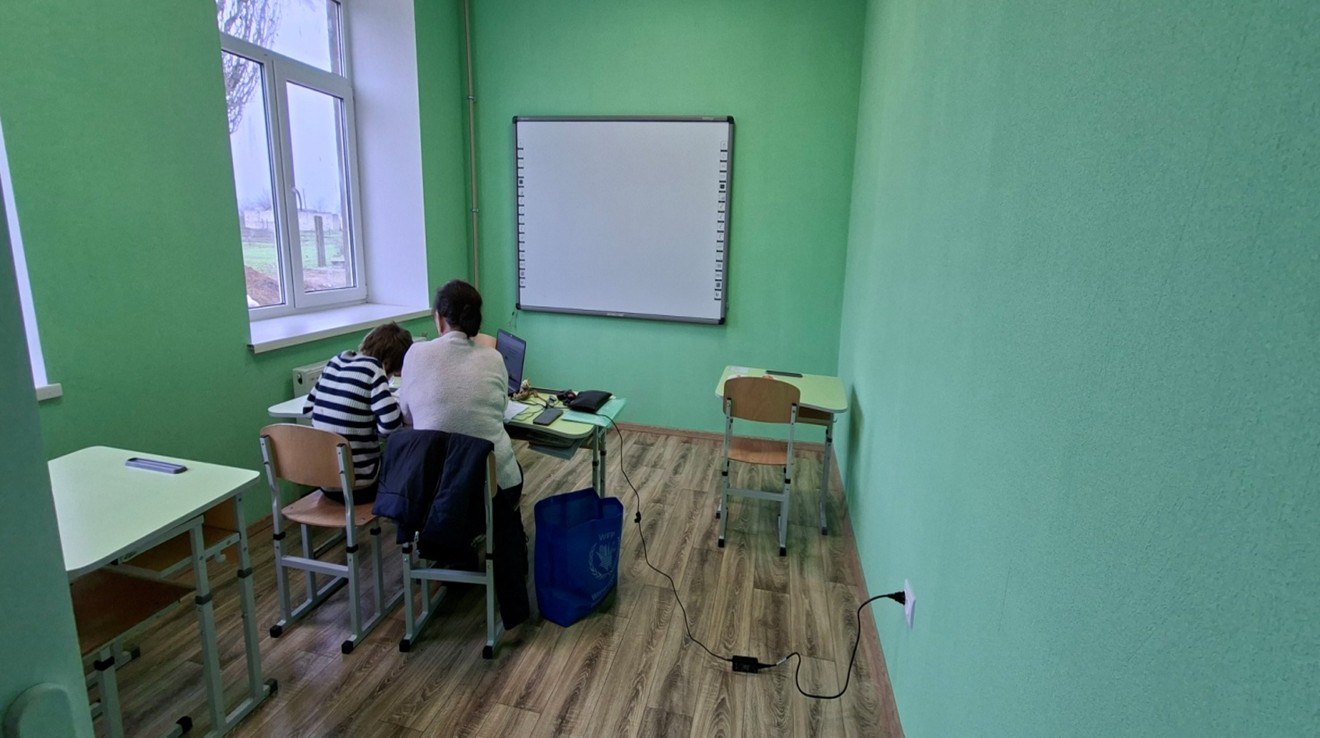 Простий клас із яскраво-зеленими стінами та дерев'яною підлогою; двоє людей сидять за столом біля великого вікна, працюючи за ноутбуком, поруч висить біла дошка.