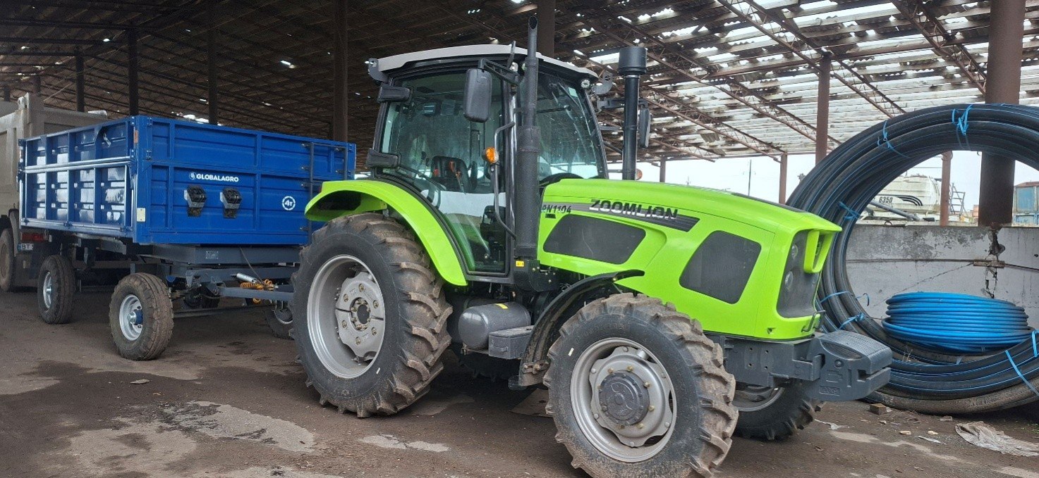 Трактор Zoomlion яскраво-зеленого кольору, причеплений до синього сільськогосподарського причепа, стоїть у приміщенні з металевими конструкціями.